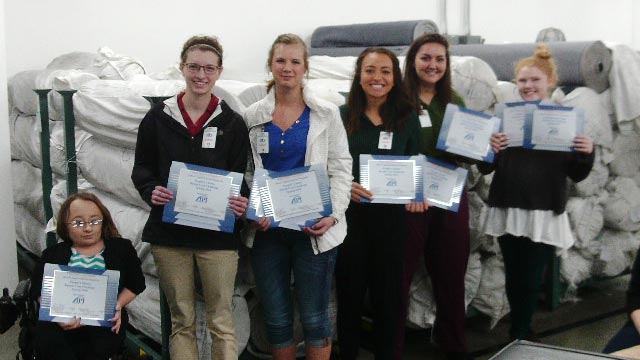 6 students standing in a line in front of large rolls of fabric, each holding their certificates.