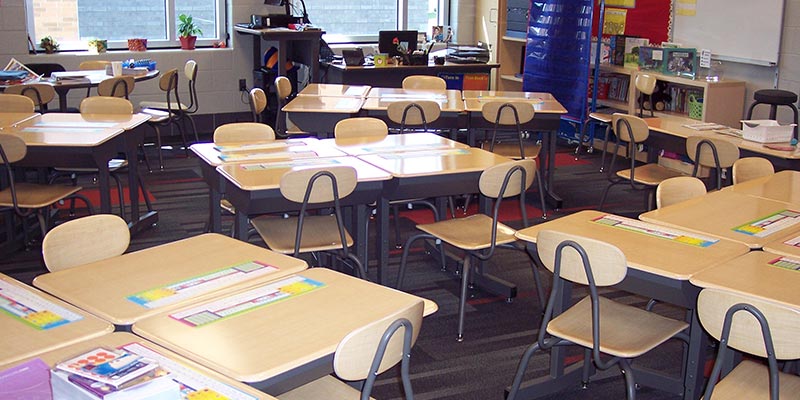 An organized elementary school classroom filled with rows of light wood student desks and matching wooden chairs, with an educator's desk and learning materials in the background.