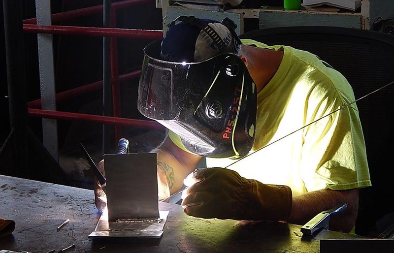 A welder wearing a protective mask and heavy gloves works on a metal piece at a workbench, with bright light from the weld illuminating the workspace.