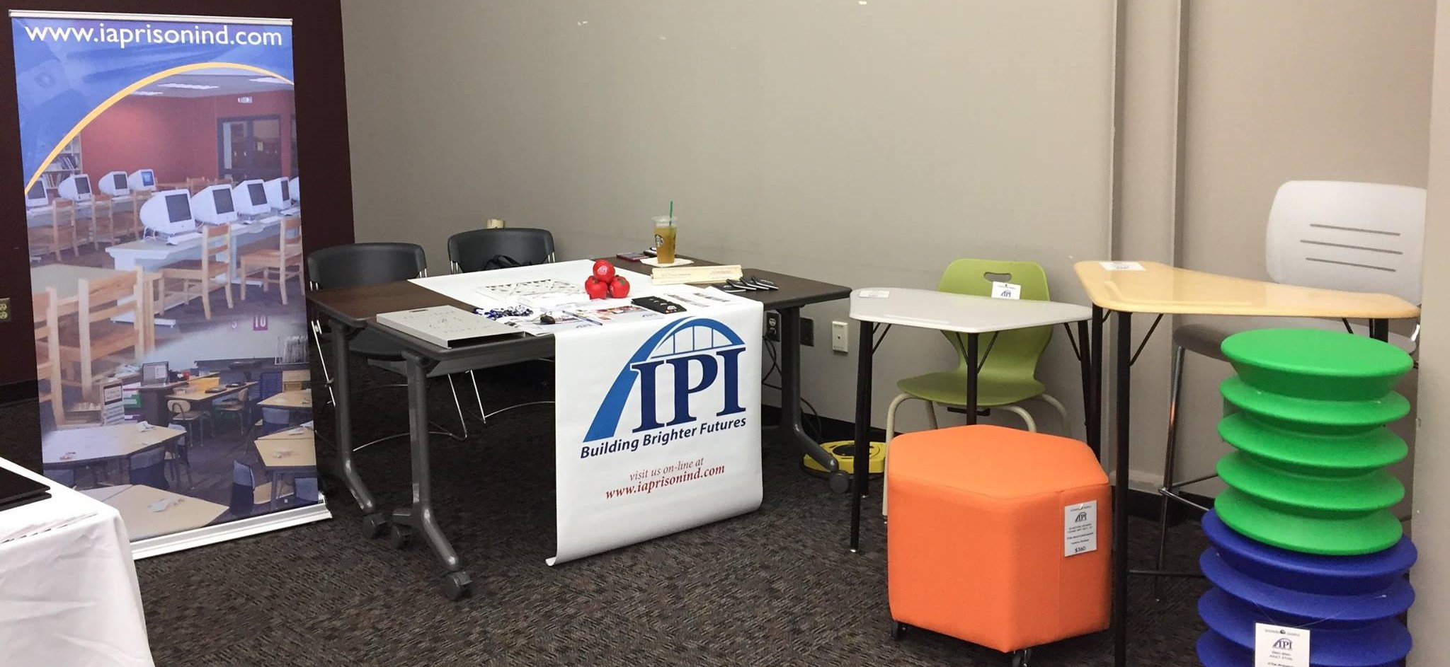 An IPI trade show booth displaying school and office furniture, featuring a banner with the IPI logo draped over a table, an orange hexagonal ottoman, colorful modular stools, student desks, and a vertical banner on the left.