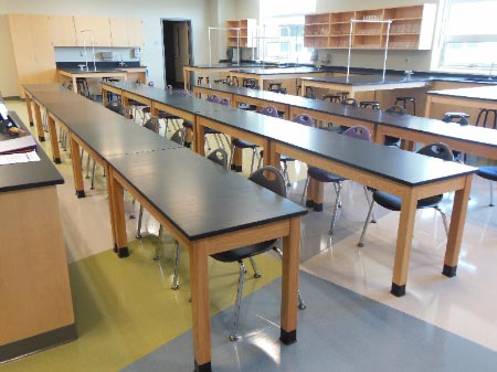 An empty school science laboratory room featuring long wooden lab tables with black tops arranged in rows, with matching cabinetry and lab workstations lining the walls.