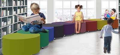 Children playing and reading on a long, winding line of colorful modular ottoman stools from the In Motion line inside a modern library setting.