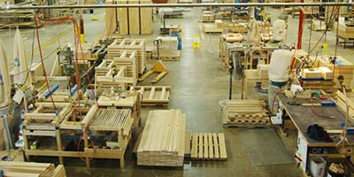 An organized industrial woodworking shop and manufacturing floor featuring organized stacks of lumber, wooden pallets, workbenches, equipment, and clearly designated walkways.