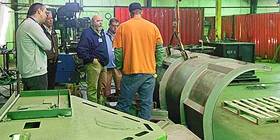 Members of the IPI sales team standing besides an unpainted snow plow on the Rockwell city plant floor listening to an incarcerated associate in an orange shirt.