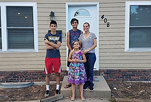 A smiling family of four—a mother, two teenage boys, and a young girl—stands proudly on the concrete front steps outside their home with tan siding and house number 408.