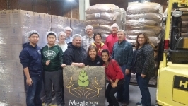 Members of IPI's leadership team standing with Meals from the Heartland volunteers in front of a brown Meals from the Heartland and boxes of food products.