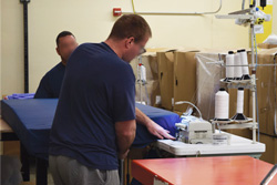 Two Fort Madison associates assembling a mattress , with one worker guiding a dark blue fabric mattress cover through an industrial sewing machine setup with large spools of white thread.