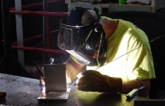 A welder wearing a protective mask and heavy gloves works on a metal piece at a workbench, with bright light from the weld illuminating the workspace.