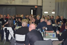 A large audience of staff seated at round tables inside a gymnasium, listening attentively to a presentation during a training conference.