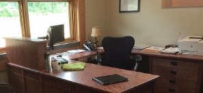 An empty office workstation featuring a large wooden wrap-around desk, a black mesh ergonomic chair, a computer monitor by a window, a telephone, a printer, and paperwork.