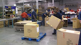 Incarcerated men in blue and orange t-shirts assembling large cardboard boxes, packing them, and placing them on blue wooden pallets.