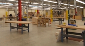 A large, open industrial area in IPI's Fort Madison woodworking shop with wooden workbenches, tools, machinery, and overhead ventilation systems under bright fluorescent lights.