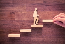 A wooden cutout of a person climbing a staircase made of small wooden blocks on a wood-grain surface, with a hand placing the next step ahead to symbolize career growth or progression.